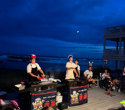 Evening beachfront hibachi show on a wooden deck — chefs cooking on portable griddles while a festive crowd in party hats watches beneath a deep blue sky and moon over the ocean.