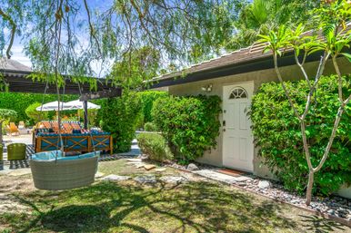 Sunny residential backyard patio with hanging wicker swing, shaded pergola over wooden lounge seating and umbrella, white entry door framed by lush green hedges