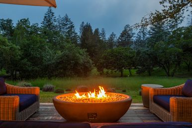 Cozy backyard gas fire pit on a wooden deck flanked by wicker patio chairs, glowing at dusk with a lush forest and meadow view