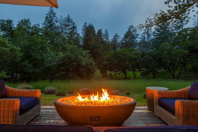 Cozy backyard gas fire pit on a wooden deck flanked by wicker patio chairs, glowing at dusk with a lush forest and meadow view