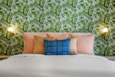 Cozy modern bedroom with tropical green leaf wallpaper, blush and tan pillows, a blue patterned accent cushion, white bedding and brass globe wall sconces