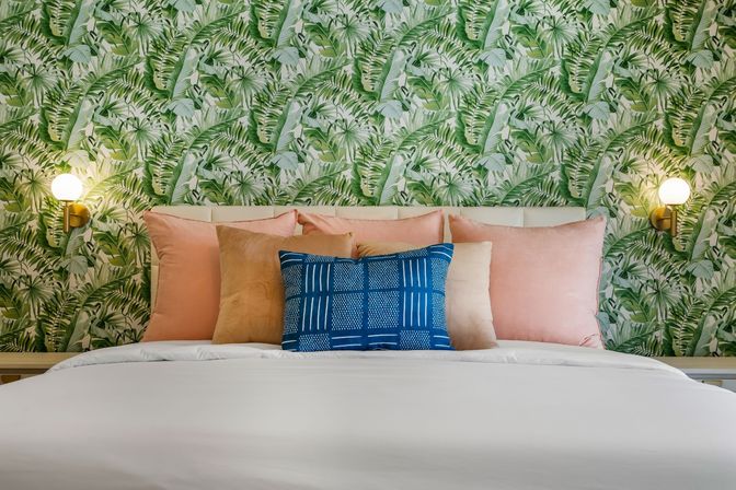 Cozy modern bedroom with tropical green leaf wallpaper, blush and tan pillows, a blue patterned accent cushion, white bedding and brass globe wall sconces
