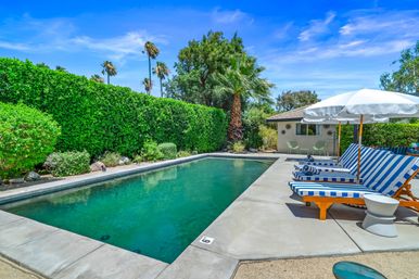 Resort-style backyard rectangular pool with palm trees, tall green privacy hedge, and blue-and-white striped loungers under white umbrellas on a sunny day