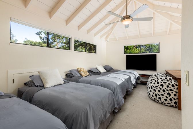Bright vaulted-ceiling guest bedroom with four gray twin beds, ceiling fan, wall-mounted TV, cow-print bean bag and treetop windows