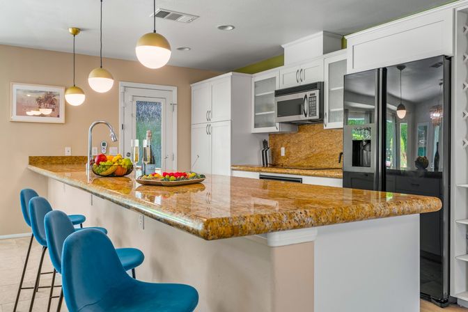 Sunny modern open-concept kitchen with a gleaming golden granite breakfast bar, three teal bar stools, globe pendant lights, white cabinets, stainless appliances, and a fruit bowl with champagne on the counter.