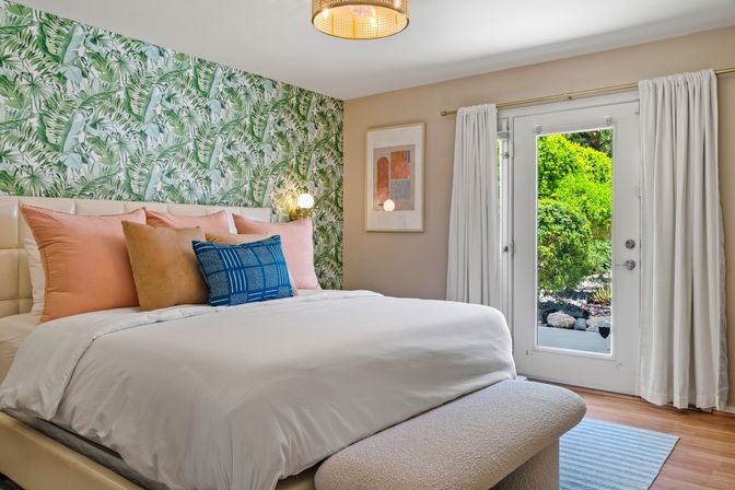 Cozy modern bedroom with tropical palm-pattern wallpaper, white bedding topped with peach and blue accent pillows, upholstered bench and glass patio doors opening to a lush garden view.