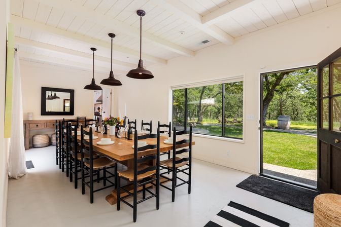 Sunlit modern farmhouse dining room with a long wooden table, twelve black ladder-back chairs, three industrial pendant lights, large window and open door leading to an oak‑shaded grassy backyard.