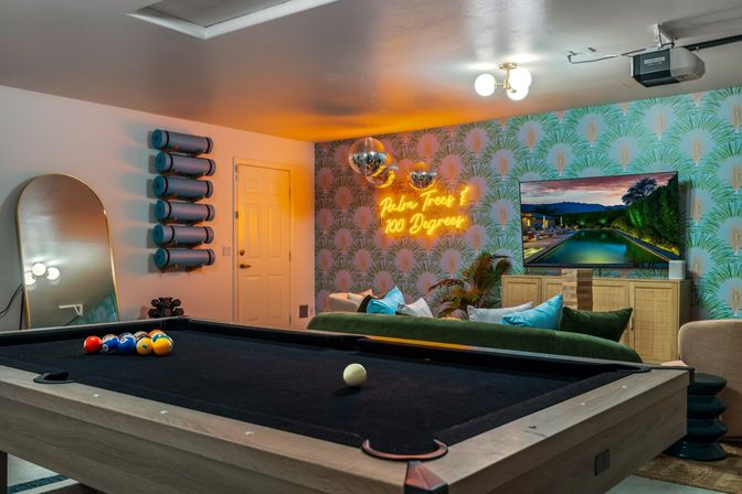 Cozy modern game room with a black-felt pool table, neon “Palm Trees 100 Degrees” sign, tropical wallpaper, disco balls, wall TV and green velvet sofa.