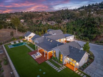 Aerial view of a modern luxury hilltop home at sunset — lit open-air deck, manicured lawn, rectangular pool, multiple gabled roofs and long driveway surrounded by wooded hills.