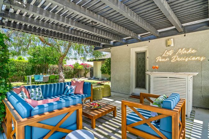 Sunny backyard patio under a slatted pergola with blue-cushioned wooden lounge seating, coffee table with snacks, small white bar and neon sign reading "Let's Make Pour Decisions"