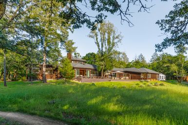 Ranch-style wooden country home with large deck and outdoor seating, surrounded by oak trees and a sunlit grassy meadow