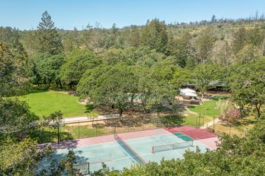 Aerial view of multi-sport courts with nets beside a sunlit green lawn, picnic tables and umbrella‑shaded patio tucked among oak trees on a wooded hillside.