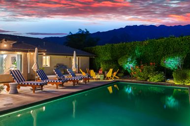 Vibrant backyard pool at sunset with turquoise water reflecting a pink-orange sky, striped lounge chairs and umbrellas on the deck, bright yellow Adirondack chairs, manicured hedge and mountain silhouette in the background.