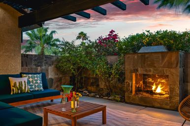 Cozy outdoor patio at sunset with a lit stone fireplace, teal cushioned seating, wooden coffee table with wine and glasses, and palm trees and flowering shrubs under a colorful sky.