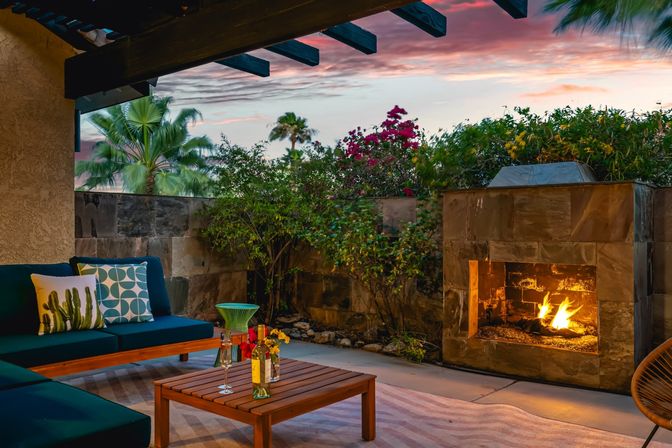 Cozy outdoor patio at sunset with a lit stone fireplace, teal cushioned seating, wooden coffee table with wine and glasses, and palm trees and flowering shrubs under a colorful sky.