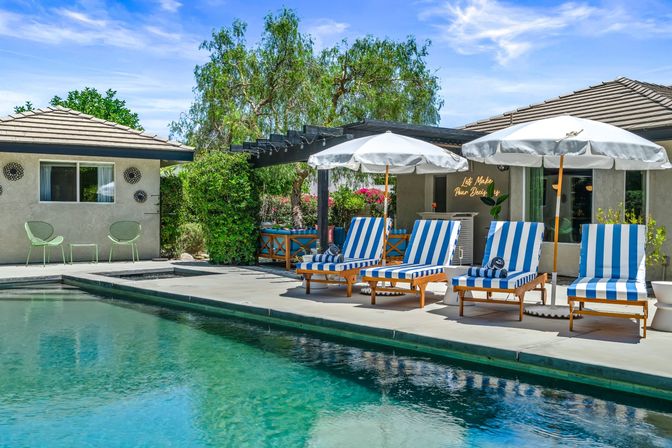 Sunny backyard swimming pool and poolside patio with blue-and-white striped lounge chairs, white umbrellas, pergola and lush greenery — inviting resort-style scene.