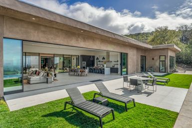 Modern hillside luxury home with floor-to-ceiling sliding glass wall opening to a patio and green lawn, sun loungers, outdoor dining table, and open-plan living room and kitchen under a bright cloudy sky.