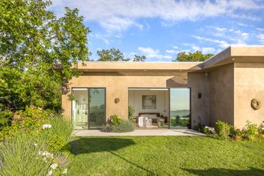 Sunlit modern stucco house with wide sliding glass doors revealing a cozy bedroom and seating area, framed by a lush green lawn, landscaped flower beds, and a leafy tree under a blue sky.