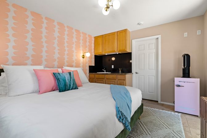 Bright cozy studio bedroom with a large bed in white linens, peach patterned accent wall, pink and teal throw pillows, small kitchenette with wooden cabinets and black backsplash, and a retro pink mini fridge.