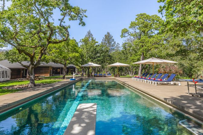 Sunlit outdoor rectangular pool at a countryside retreat with a diving board, blue lounge chairs topped with red-striped towels under large umbrellas, shady oak trees and rustic cabins in the background