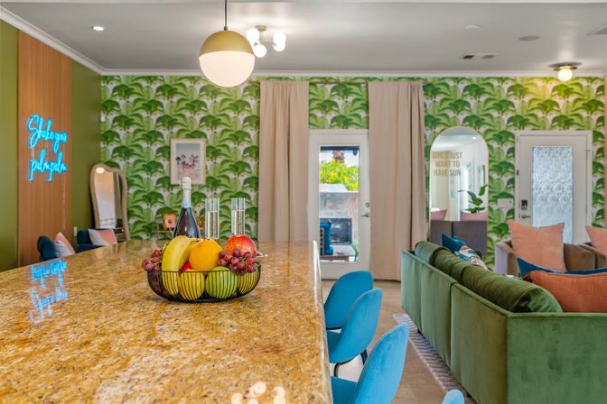 Open-plan tropical-themed living room and kitchen with a granite island topped by a fruit bowl and champagne, teal bar stools, palm-leaf wallpaper, and green velvet sofa.