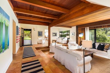 Sunlit open living room with exposed wooden beams, white slipcovered sofas, terracotta tile and striped runners, and large windows overlooking a green yard.