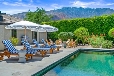 Sun-soaked backyard pool and concrete patio with blue-and-white striped lounge chairs, white umbrellas, wooden seating and lush hedges, framed by a rugged mountain backdrop