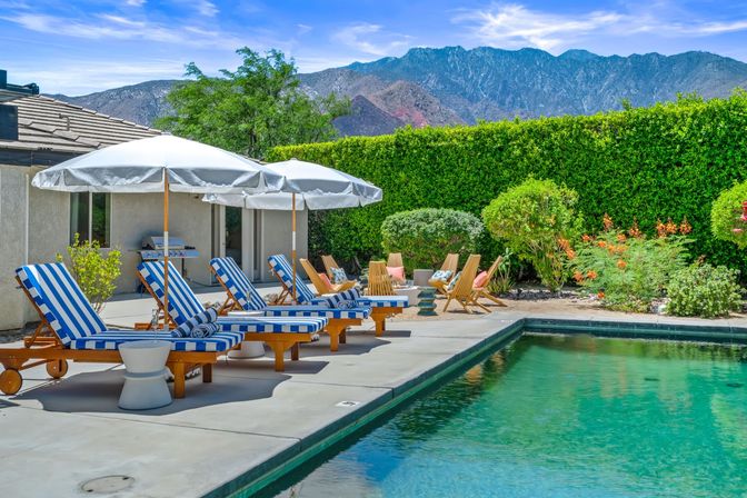 Sun-soaked backyard pool and concrete patio with blue-and-white striped lounge chairs, white umbrellas, wooden seating and lush hedges, framed by a rugged mountain backdrop