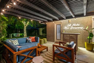 Cozy backyard patio lounge at night with blue cushioned wooden sofa and chairs on a striped rug, string lights overhead, tree wrapped in fairy lights, and a neon wall sign reading "Let's Make Pour Decisions" above a small outdoor bar.