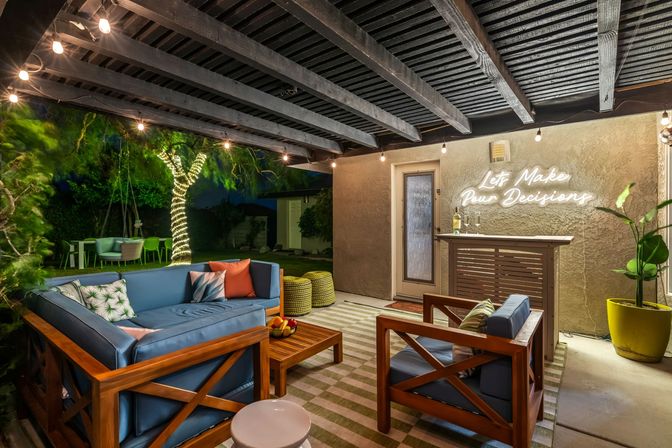 Cozy backyard patio lounge at night with blue cushioned wooden sofa and chairs on a striped rug, string lights overhead, tree wrapped in fairy lights, and a neon wall sign reading "Let's Make Pour Decisions" above a small outdoor bar.