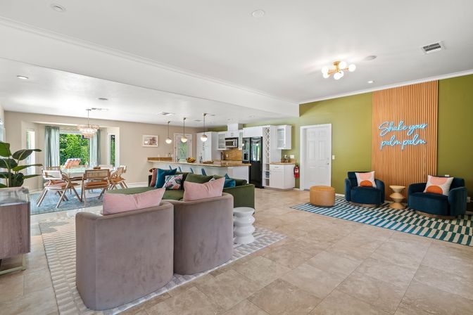 Sunlit modern open-concept living room and kitchen with tiled floor, green accent wall featuring a neon “Shake your palm palm” sign, velvet armchairs, sectional sofa with pastel pillows, dining table by sliding glass doors, and a kitchen island with pendant lights.