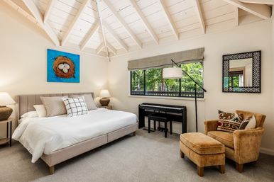 Bright, airy bedroom with white vaulted wood ceiling, king bed with neutral linens, blue nest artwork, digital piano under a window with tree-lined view, and a cozy velvet armchair with ottoman.