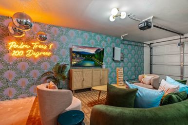 Tropical-themed converted garage lounge with neon sign reading "Palm Trees & 100 Degrees", mirrored disco balls, palm-print wallpaper, curved sofas and armchairs around a coffee table and wall-mounted TV, garage door visible.
