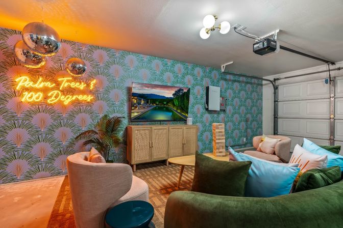 Tropical-themed converted garage lounge with neon sign reading "Palm Trees & 100 Degrees", mirrored disco balls, palm-print wallpaper, curved sofas and armchairs around a coffee table and wall-mounted TV, garage door visible.