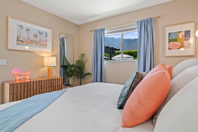 Bright modern bedroom with white bed, coral pillows and light-blue runner, wooden dresser with neon dachshund light and lamp, potted palm, tall mirror and blue curtains framing a window with a mountain view.