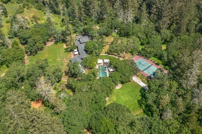 Aerial drone view of a secluded country estate tucked in leafy woodland, showing a main house, rectangular swimming pool, adjacent tennis courts and manicured green lawns.