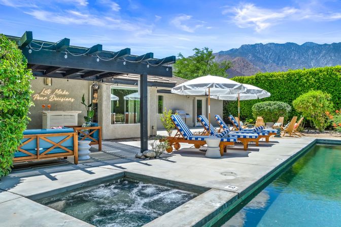 Sunny backyard oasis with bubbling spa and long pool, blue-and-white striped lounge chairs under white umbrellas, a wooden pergola with string lights and neon sign, manicured hedges and mountain backdrop.