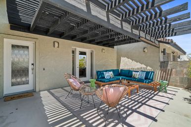 Sunny modern backyard patio with black slatted pergola casting striped shadows over a teal L-shaped sectional, wooden coffee table, pink-cushioned wicker chairs and green side tables on a chevron rug.