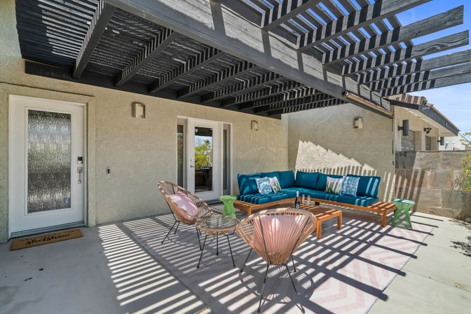 Sunny modern backyard patio with black slatted pergola casting striped shadows over a teal L-shaped sectional, wooden coffee table, pink-cushioned wicker chairs and green side tables on a chevron rug.