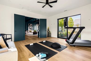 Sunlit modern home gym with treadmill by large sliding glass door, two black yoga mats with towels, balance trainer and foam blocks on light oak floors, teal double doors open to a cozy living room