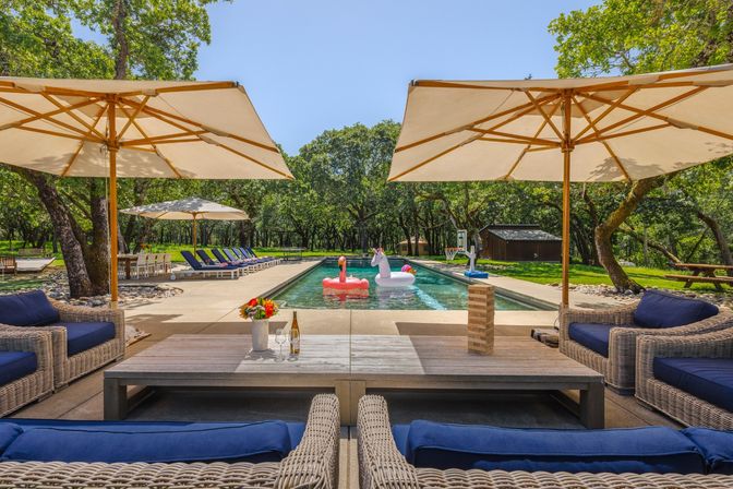 Oak-shaded backyard lap pool with inflatable flamingo and unicorn, blue-cushioned wicker sofas, large beige umbrellas, lined lounge chairs and picnic table under a clear summer sky.
