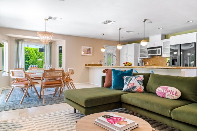 Open-concept sunny living room and kitchen in a suburban home — green velvet sectional with tropical pillows, rattan dining nook, breakfast bar with pendant lights and sliding glass door to a green backyard.