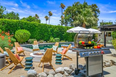 Sunny backyard pool patio with stainless steel gas grill topped with colorful bell peppers, round fire pit surrounded by wooden lounge chairs, poolside umbrella, tall palm trees and dense green hedge