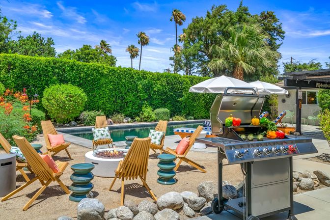 Sunny backyard pool patio with stainless steel gas grill topped with colorful bell peppers, round fire pit surrounded by wooden lounge chairs, poolside umbrella, tall palm trees and dense green hedge