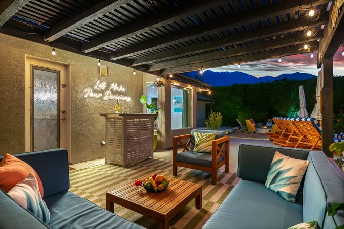 Mountain-view backyard patio at twilight — covered outdoor lounge with blue cushioned sofas, wooden coffee table with fruit bowl, neon “Let’s Make Pour Decisions” sign, string lights and poolside loungers