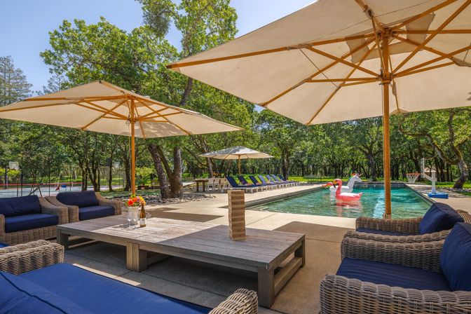 Resort-style backyard pool deck with wicker lounge seating and navy cushions, large patio umbrellas, row of sun loungers, inflatable flamingo and unicorn in the pool, surrounded by oak trees and a poolside basketball hoop.