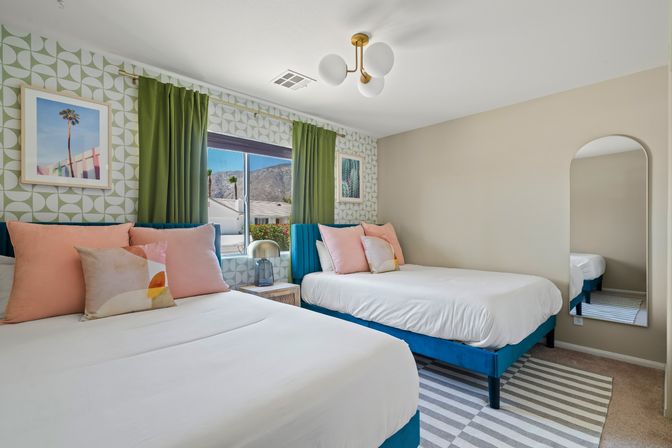Bright midcentury-modern guest bedroom with two white beds on teal frames, peach throw pillows, geometric green wallpaper, green curtains framing a mountain-view window, globe ceiling light and arched wall mirror.