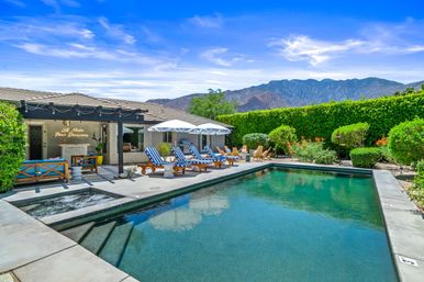 Resort-style backyard with rectangular swimming pool and built-in spa, striped sun loungers and white umbrellas on a concrete patio, covered seating with string lights, manicured hedges, flowering shrubs and mountain backdrop under a bright blue sky.