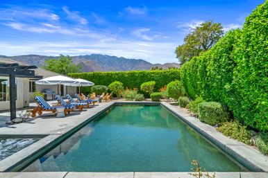 Sunny backyard rectangular pool with blue-striped lounge chairs and umbrellas, tall privacy hedges, mountain backdrop and clear blue sky.