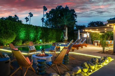 Cozy sunset poolside patio with palm trees, string lights and loungers — wooden chairs arranged around a glowing fire pit, illuminated pool and landscaped hedges.